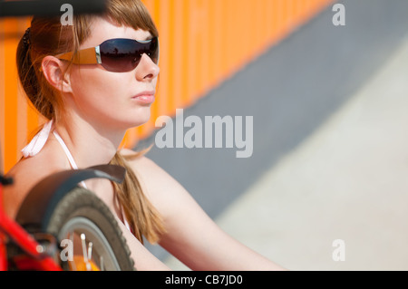 Bella giovane donna seduta vicino alla parete, indossando occhiali da sole. Frammenti della sua bicicletta di fronte a lei. Foto Stock