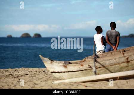 Due giovani uomini contemplativi chat della spiaggia vicino a un malgascio canoa outrigger, con piccole isole in background. Foto Stock