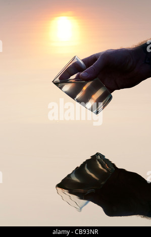 Mano raccogliendo l'acqua in un bicchiere in un lago ancora a sunrise in India. Silhouette Foto Stock