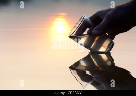 Mano raccogliendo l'acqua in un bicchiere in un lago ancora a sunrise in India. Silhouette Foto Stock