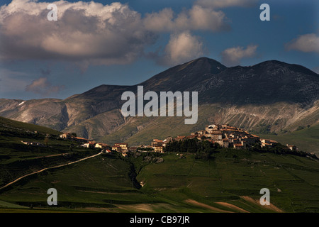 Il borgo di Castelluccio arroccato sopra il piano Grande con le montagne del Parco Nazionale dei Monti Sibillini, Umbria, Italia Foto Stock