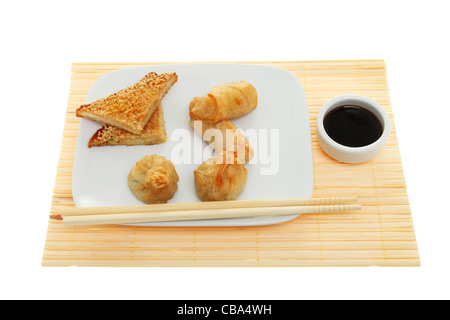 Spuntini cinesi, spring roll, i wonton e toast di gamberi su una piastra con bacchette e salsa di soia in un ramekin su un bambù matt Foto Stock