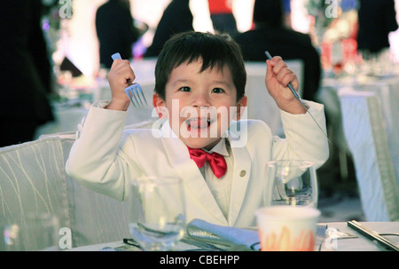 Eurasian boy in smoking bianco tuta durante un banchetto di nozze pronto a mangiare. ha fame tenendo la sua forchetta e coltello in aria Foto Stock