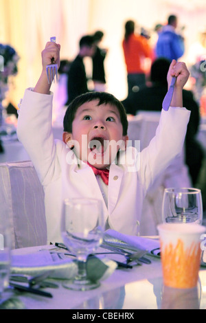 Eurasian boy in smoking bianco tuta durante un banchetto di nozze pronto a mangiare. ha fame tenendo la sua forchetta e coltello in aria Foto Stock