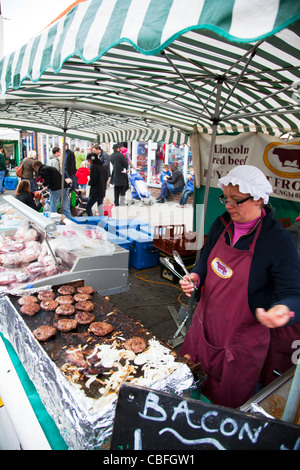 Louth mercato Vittoriano, Lincolnshire, Inghilterra donna con alette la cottura di hamburger, cipolla e pancetta per i clienti Foto Stock