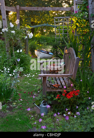 panca da giardino e rose sotto perigola in giardino floreale con attrezzi da giardinaggio tradizionali barca a remi e fiume dietro al tramonto Foto Stock