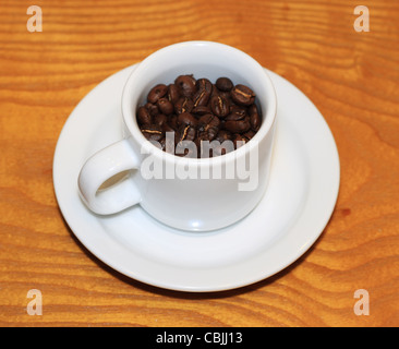Di piccole dimensioni e di colore bianco tazza di chicchi di caffè tostati con piattino su uno sfondo di legno Foto Stock