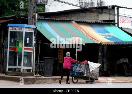 Una donna che vive in condizioni di povertà sta spingendo un cestino della spesa su una strada di città di Krabi, in Thailandia. Foto Stock