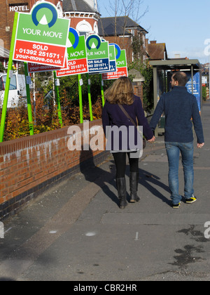 Coppia giovane camminando per strada a guardare una casa in vendita e casa venduta segni su una strada nel centro di Exeter Regno Unito Foto Stock