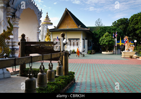 Monaco buddista in tunica arancione cammina con carriola a tempio in Thailandia del Nord con 4 campane su una trave di legno in primo piano Foto Stock