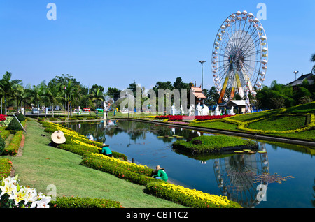 Giardinieri con enormi cappelli facendo manutenzione del paesaggio accanto al laghetto a expo in Chiang Mai Thailandia con ruota panoramica Ferris in background Foto Stock
