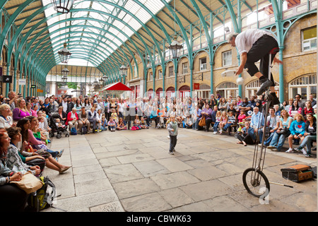 I turisti e gli amanti dello shopping e un attore di strada su una bicicletta alto con un pubblico al Covent Garden di Londra, Inghilterra. Foto Stock