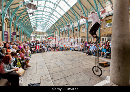 I turisti e gli amanti dello shopping e un attore di strada su una bicicletta alto con un pubblico al Covent Garden di Londra, Inghilterra. Foto Stock