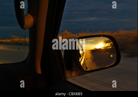 Sun discendente, visto nel retrovisore durante la guida su una strada di ghiaia nel Parco Nazionale Etosha al crepuscolo, Namibia. Foto Stock