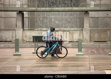 L'uomo spingendo una bicicletta di indossare una felpa con cappuccio caldo sul marciapiede davanti a cascata in ambiente urbano di Salt Lake City, Utah, Stati Uniti d'America. Tempesta di pioggia. Foto Stock