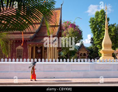 Un arancione-derubato monaco buddista cammina per la strada di fronte al Wat Sen Tempio a Luang Prabang, Laos. Foto Stock