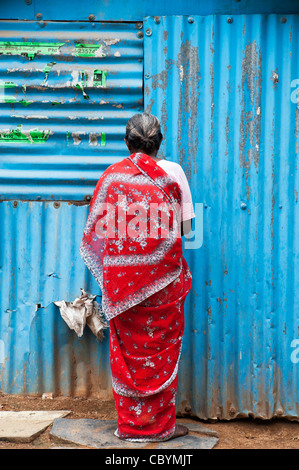 Il vecchio donna indiana in un sari rosso in piedi di fronte ad uno stagno blu shack. Andhra Pradesh, India Foto Stock