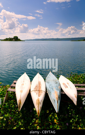 Canoe sul lago Peten Itza Flores Guatemala | FLORES, Guatemala — Quattro canoe consumate riposano con lo scafo in alto su una rastrelliera di legno sopra le canne del lago Peten Itza, con il piccolo isolotto di Radio Petén visibile in lontananza. La stazione trasmette da quell'isolotto dal 1947, e il lago stesso circonda Flores, costruita sul sito di Nojpetén, l'ultimo regno Maya indipendente, conquistato dalle forze spagnole sotto Martín de Ursúa nel 1697. Foto Stock