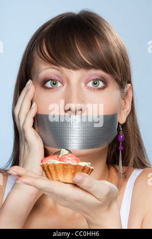 Bella ragazza intelligente fare del suo meglio per non mangiare la torta con la preoccupazione nei suoi occhi e il nastro adesivo sulla bocca Foto Stock