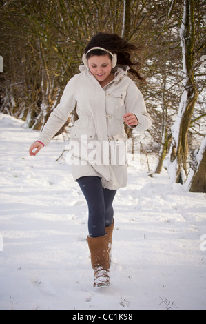 Giovane donna caucasica all'aperto che corre nella neve indossando un caldo cappotto bianco, stivali, cuffie Foto Stock