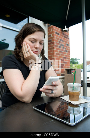 La donna caucasica guarda al suo iPhone mentre iPad si appoggia sul tavolo in un cafe' all'aperto Foto Stock