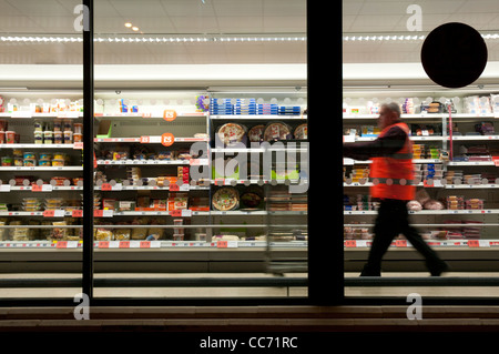 Dipendente in un giubbotto arancione spingendo un carrello giù un supermercato corsia fotografata da fuori di notte Foto Stock