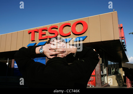 Un uomo tiene le mani dietro la testa davanti a un supermercato Tesco Foto Stock