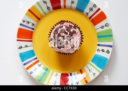 Guardando verso il basso sul cupcake alla fragola con cuore di cioccolatini a forma di spruzzata sulla sommità collocato sulla colorata gialla - piastra da sopra Foto Stock