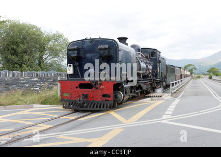 Una locomotiva a vapore con un treno passeggeri sulla Welsh Highland linea che attraversa la strada nei pressi di Porthmadog Foto Stock