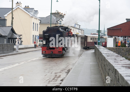 Una locomotiva a vapore con un treno passeggeri sulla Welsh Highland linea che attraversa la strada a Porthmadog Foto Stock