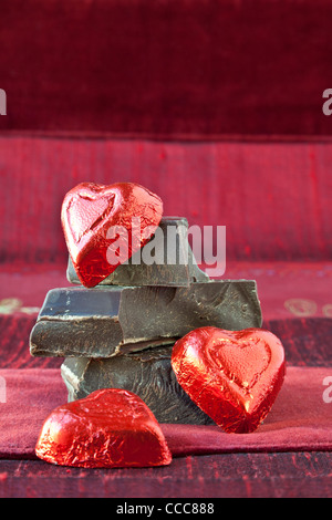 Rosso di stagnola avvolto candy cuori disposti sulla sommità della pila di spessore gourmet dark chocolate bar chunk. Rosso sfondo a trama. Foto Stock
