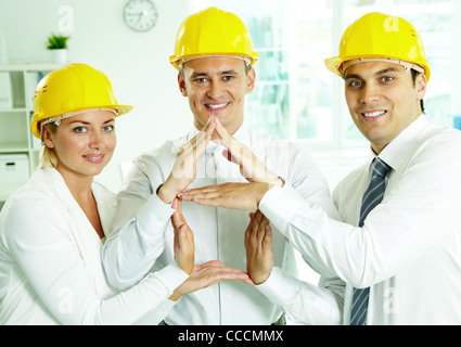 Un gruppo di architetti sorridente in caschi che mostra la mano segno di casa Foto Stock