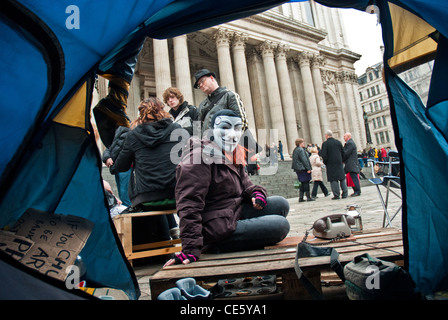 Foto scattata guardando fuori da una tenda di Occupy St Pauls con un manifestante mascherato, anche i visitatori e le colonne di St Pauls sullo sfondo Foto Stock