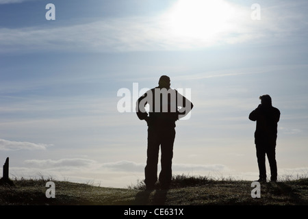 Silhouette di due uomini guardando la vista dalla cima di una collina retroilluminati da sun. Newborough Warren, Isola di Anglesey, Galles del Nord, Regno Unito. Foto Stock