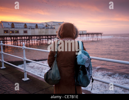 Vista posteriore di una donna che indossa un cappotto duffel guardando gli storni sono ' appollaiati sul molo al tramonto , Aberystwyth Wales UK Foto Stock