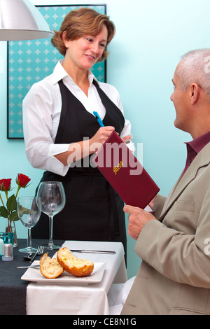 Foto di una cameriera in un ristorante prendendo un ordine alimentare da un maschio maturo chi è seduto al tavolo tenendo un menu. Foto Stock