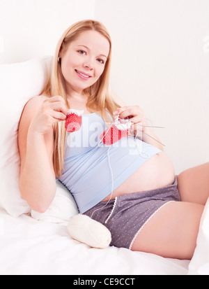 Bella giovane donna incinta maglia baby's bootees nel letto di casa sorridente Foto Stock