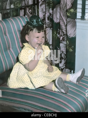 Colorati a mano vintage fotografia di 2 anno di età bambino bambina indossa una festa di compleanno hat e seduto su una sedia imbottita, 1949. Foto Stock