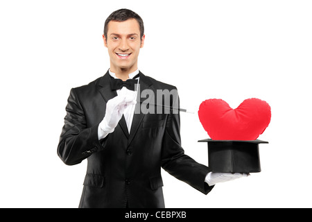 Giovane mago in possesso di una bacchetta magica e top hat con un cuore rosso a forma di cuscino isolato su sfondo bianco Foto Stock