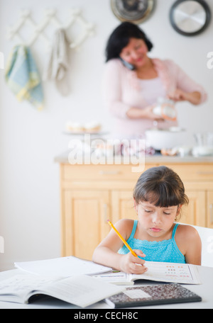 Ragazza ispanica facendo compiti a casa con la madre in background Foto Stock