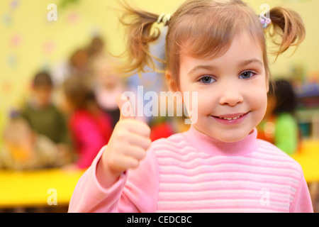 Poco sorridente ragazza in kindergarten mostra segno ok Foto Stock