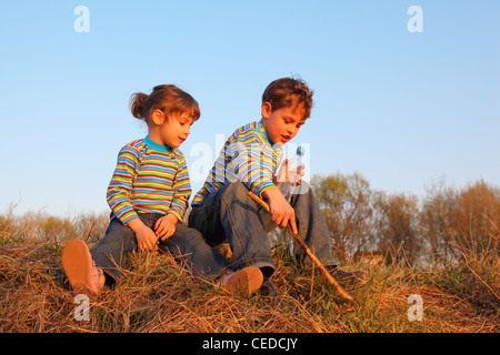 Bambina e ragazzo con bottiglia e stick in striped t-shirts sedersi su erba secca Foto Stock