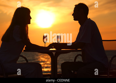 Donna e uomo di sagome su sunset sedere al tavolo con due bicchieri all'aperto, holding per le mani Foto Stock