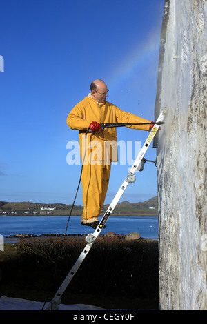 L'uomo una scaletta utilizzando la rondella a pressione per rimuovere la vernice da un bianco muro di casa Foto Stock