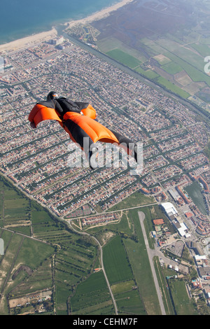 Uomo in tuta con Orange Wings è volare sopra una città turistica vicino al mare. Lui è molto felice per volare nel cielo blu. Foto Stock