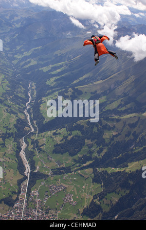 Uomo in tuta con Orange Wings è volare sopra le montagne verdi accanto al bianco delle nuvole. Foto Stock