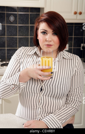 Il sovrappeso donna seduta in cucina con un bicchiere di succo di arancia. Foto Stock
