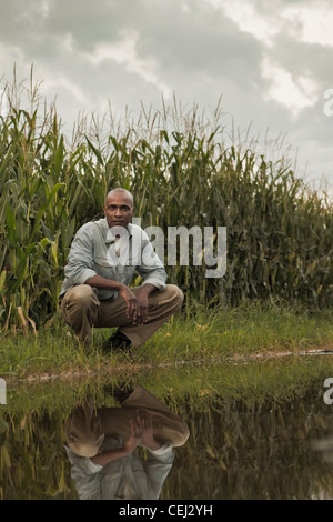 African American Farmer squatting in prossimità di acqua e le colture Foto Stock