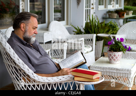 Uomo maturo di leggere la sua Bibbia mentre è seduto in un bianco sedia di vimini su un portico. Foto Stock