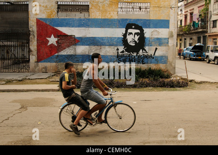 Ragazzi a piedi con la bicicletta nella parte anteriore di una bandiera cubana e iconico Che Guevara pittura murale a città di Havana, Cuba Foto Stock